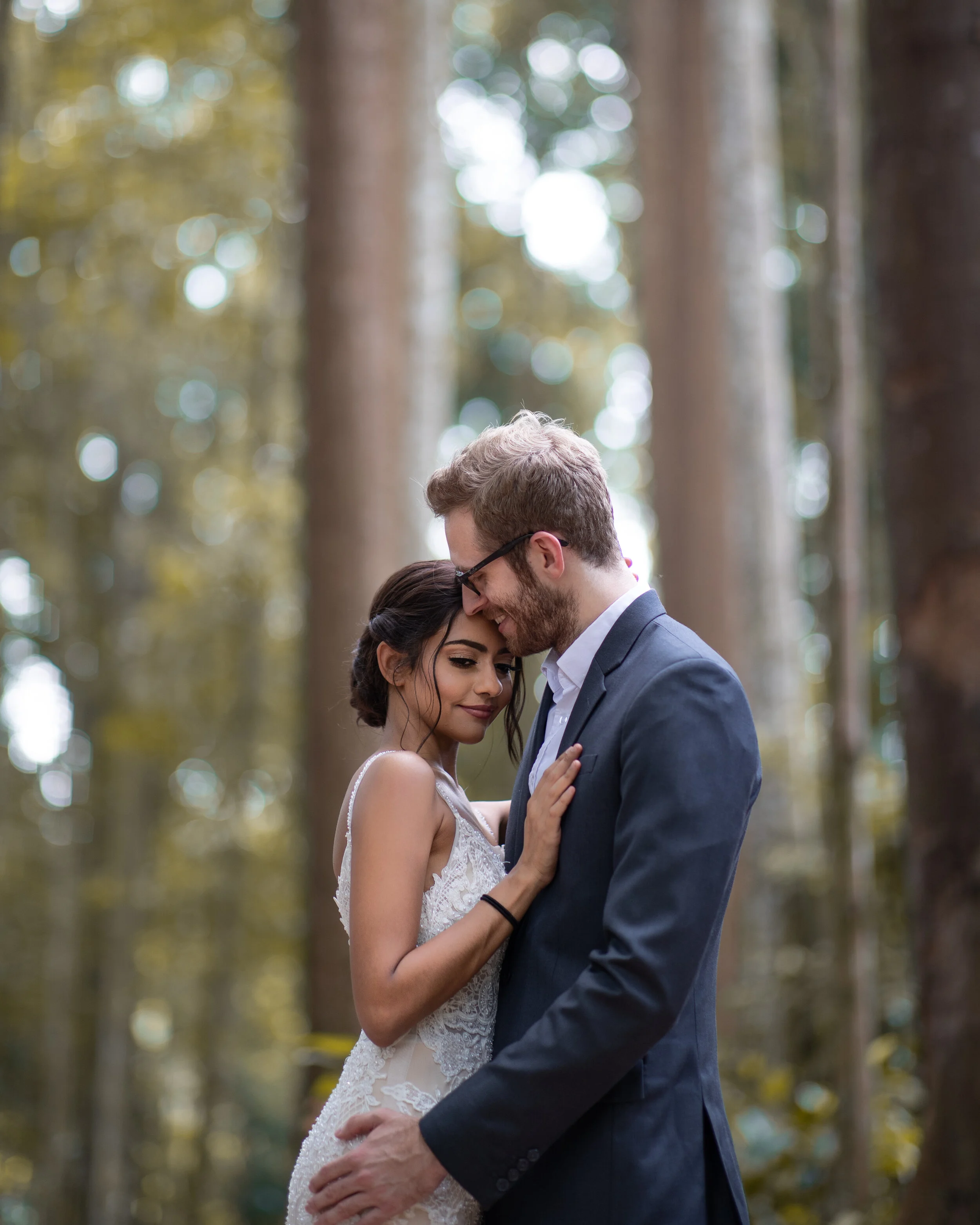 A man and woman embrace outdoors in a forest, in a close, tender pose, with trees and sunlight in the background.