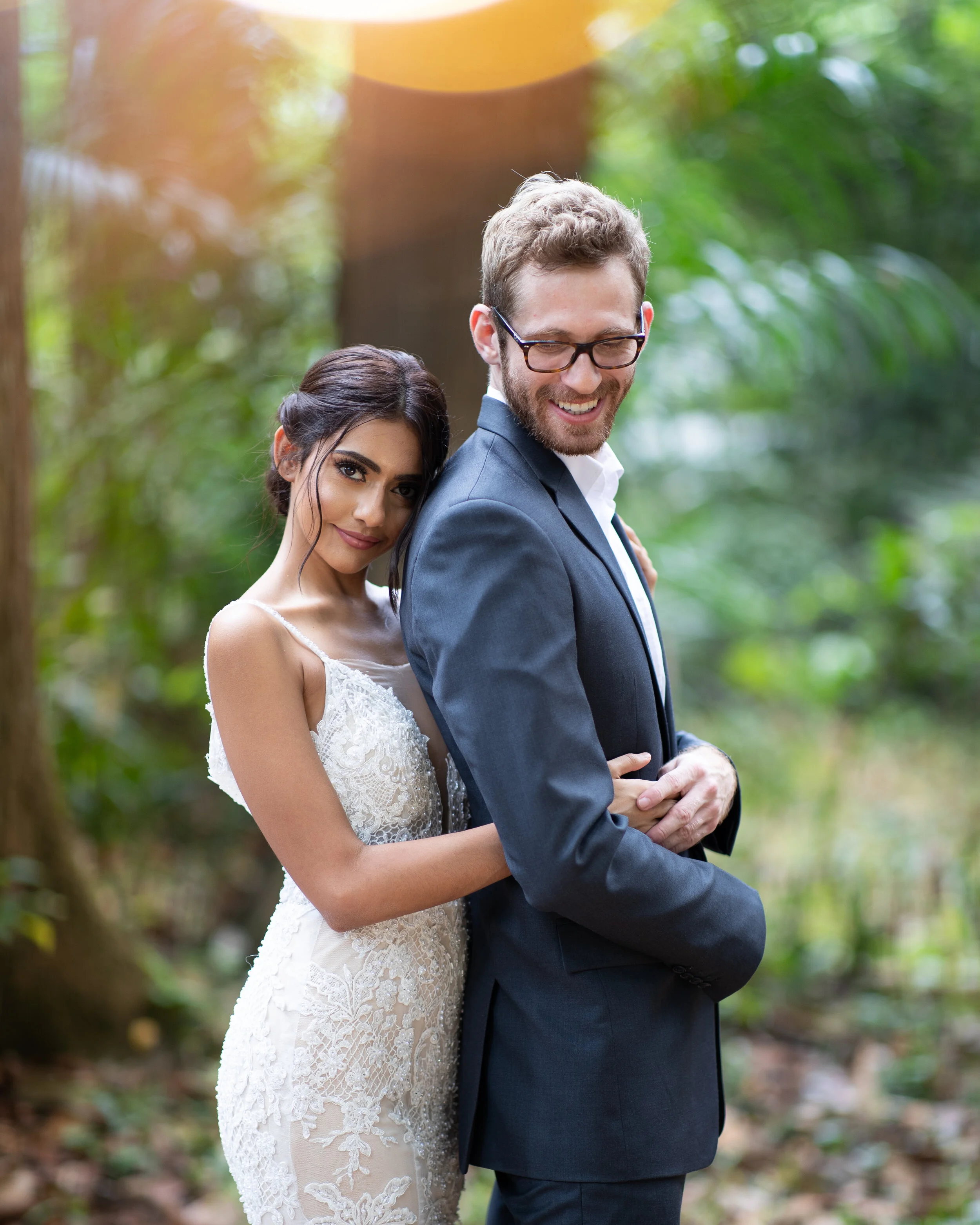 A joyful couple in wedding attire standing close, outdoors in a lush green park. The woman, with dark hair styled in an elegant updo, is leaning her head on the man's shoulder. She is wearing a detailed white lace wedding dress. The man, with short c