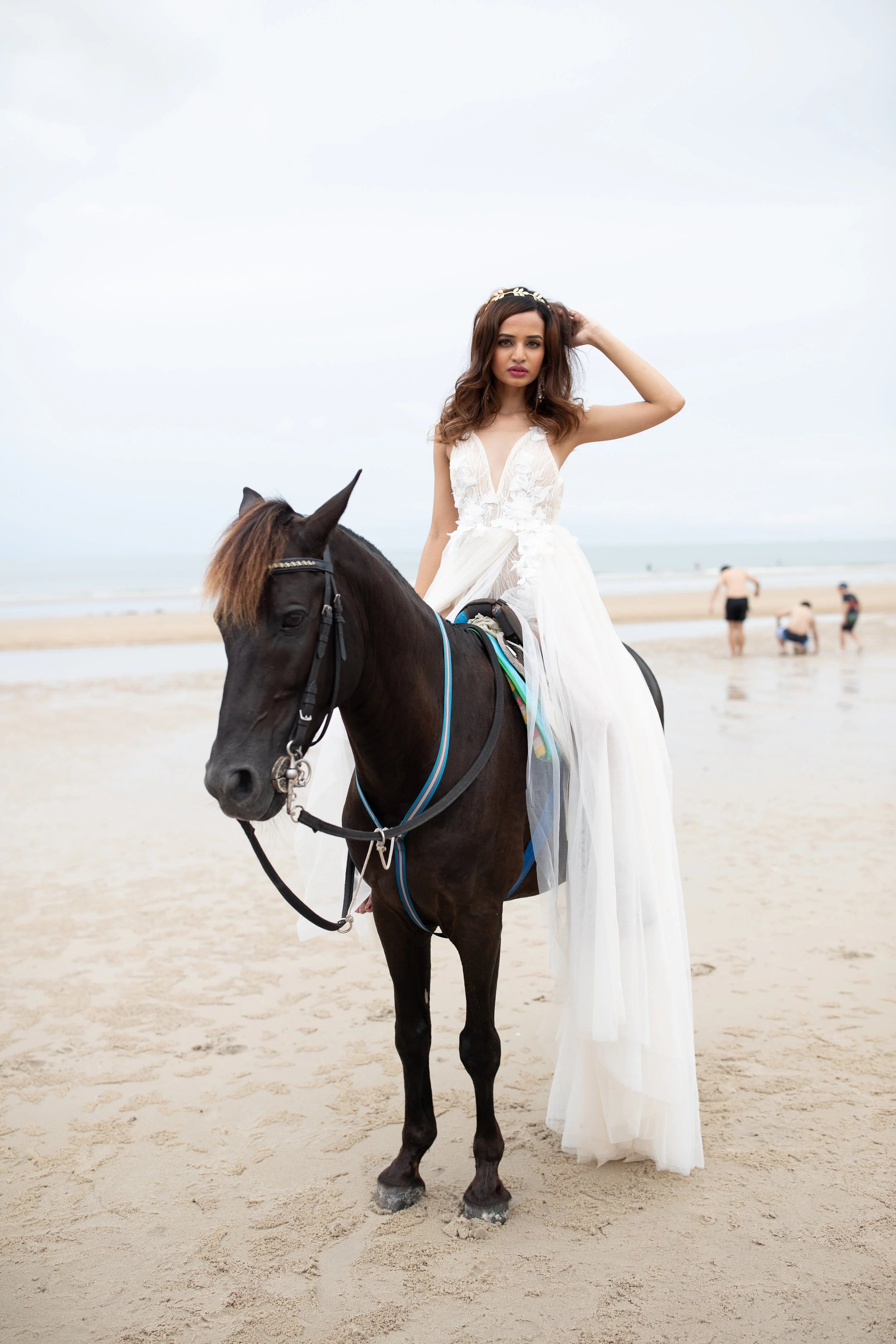 A woman dressed in a white gown riding a black horse on a beach, with a cloudy sky and people in the background.