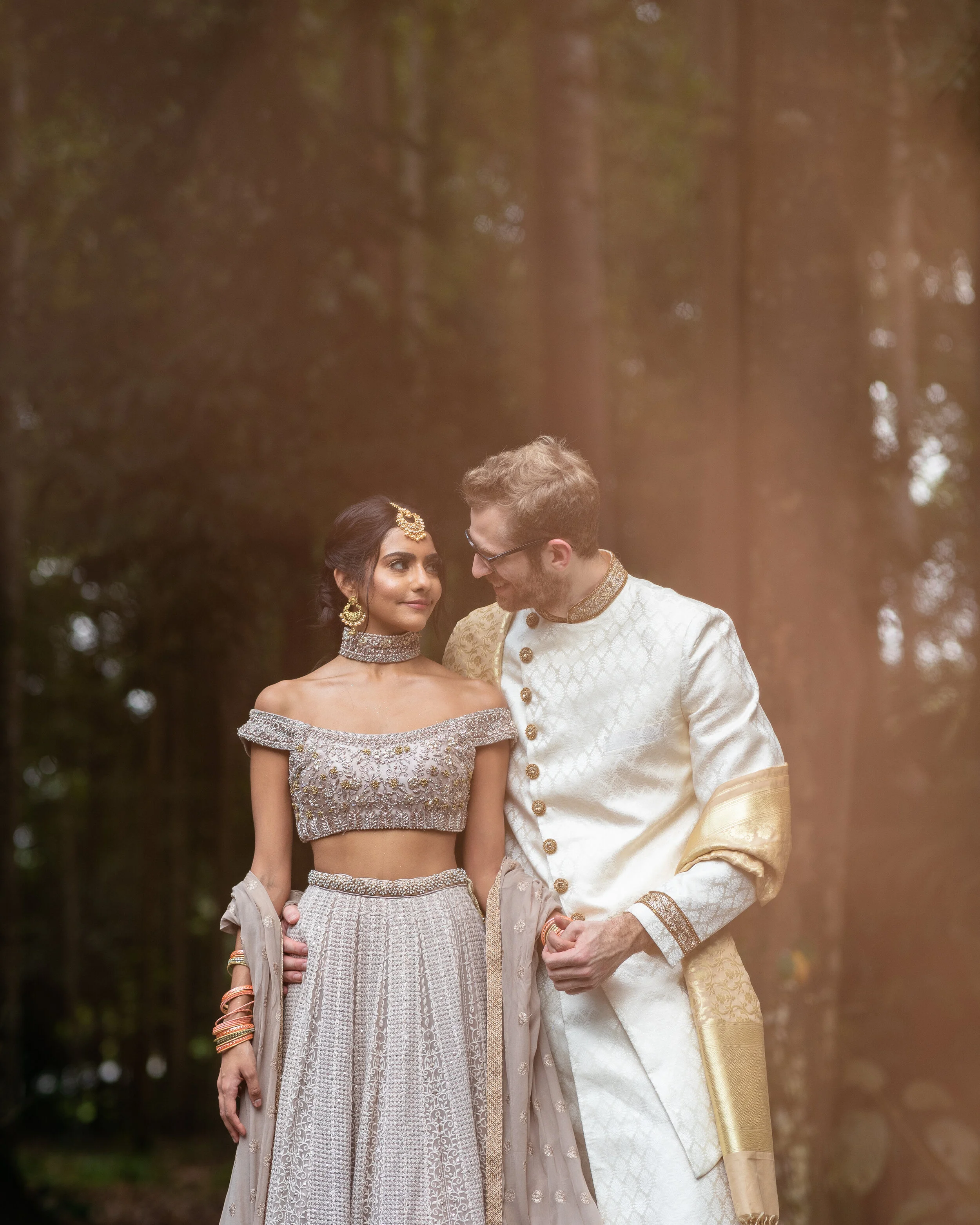 A couple dressed in traditional Indian wedding attire standing close together outdoors, surrounded by trees.