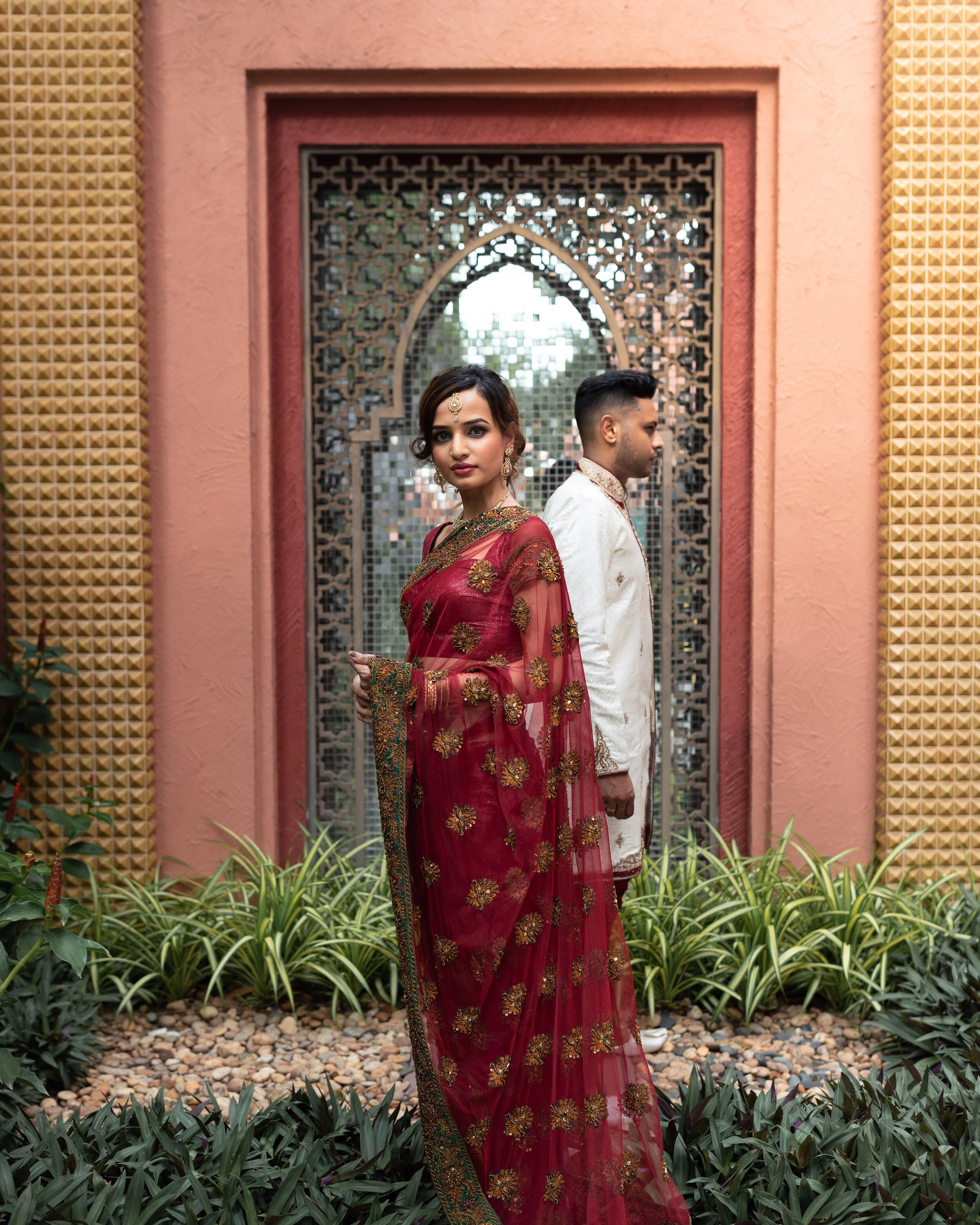 A woman and a man dressed in traditional Indian attire standing back to back in front of an ornate window with a pink and red wall, surrounded by green plants.