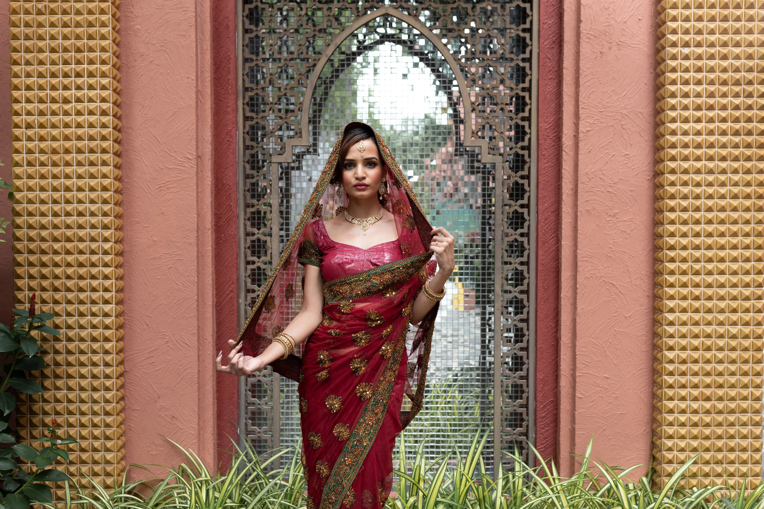 A woman dressed in a traditional red sari with gold embroidery, standing in front of an ornate metal gate framed by pink textured walls.