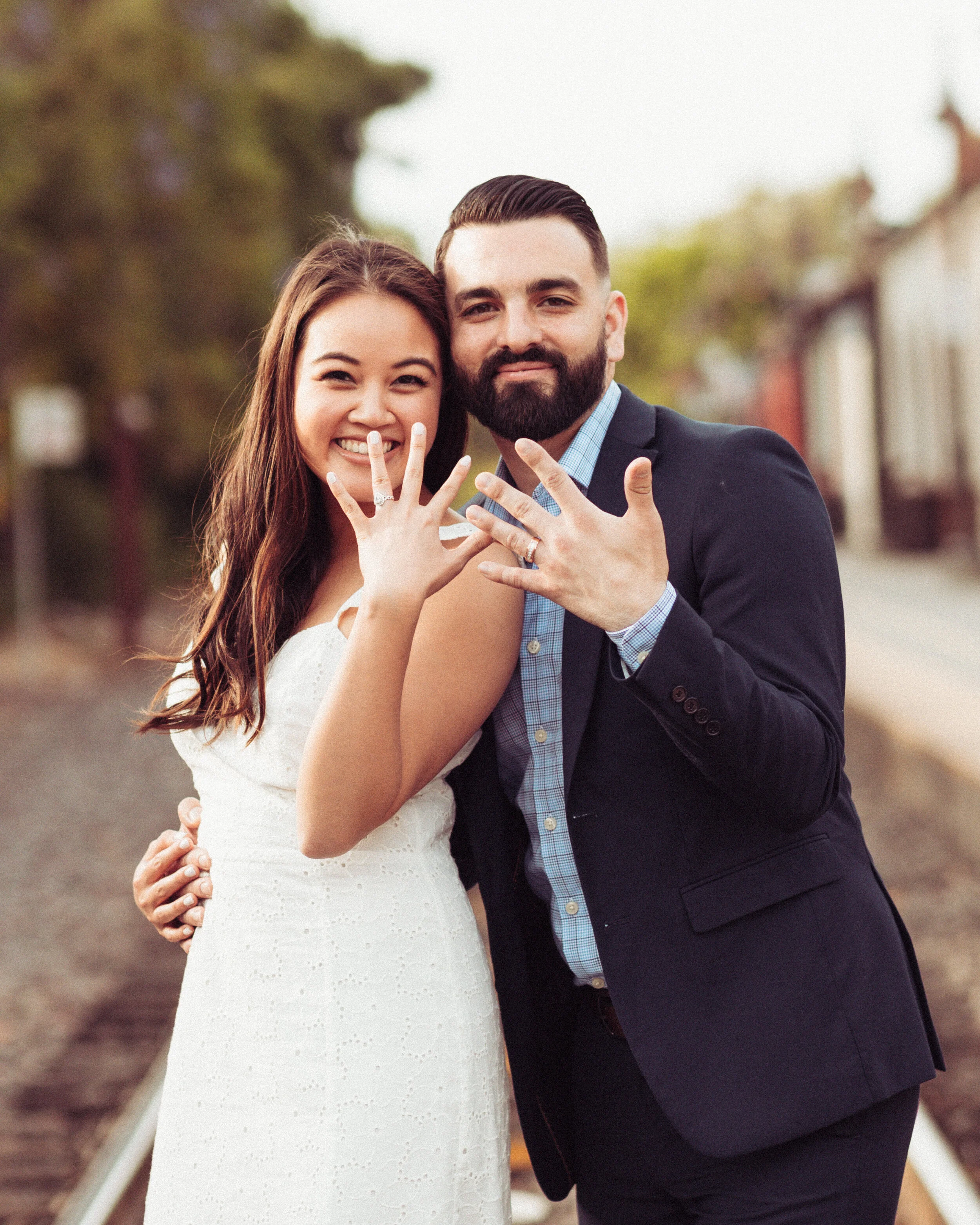 A happy couple showing their wedding rings, standing outdoors with trees and buildings in the background.