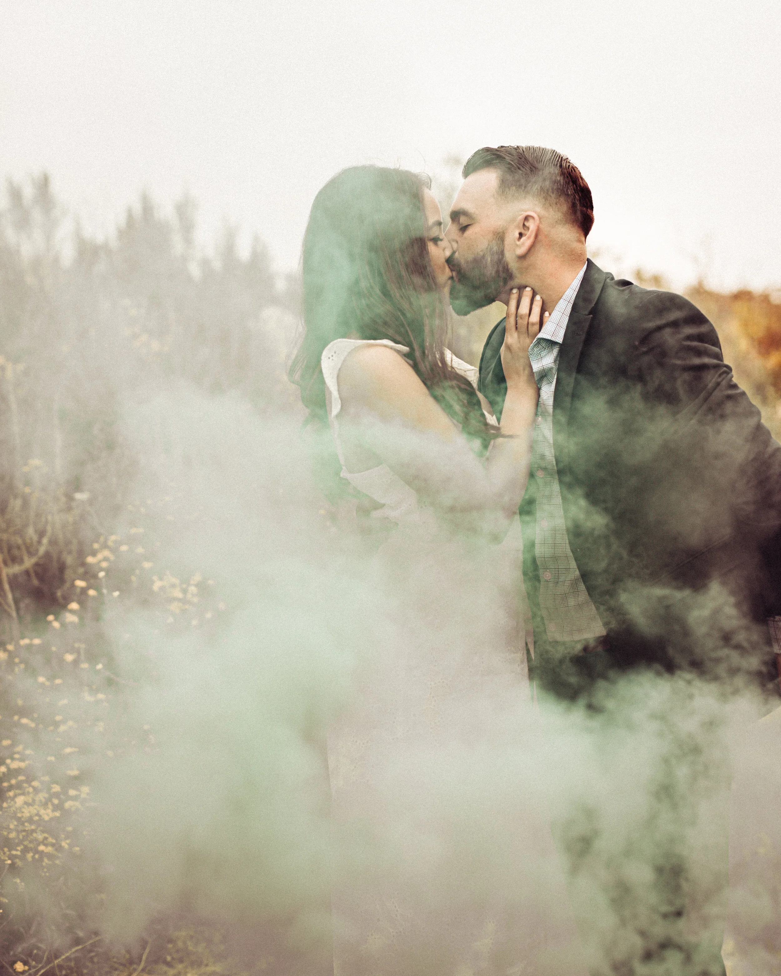 A couple sharing a kiss outdoors in a foggy setting with trees in the background.