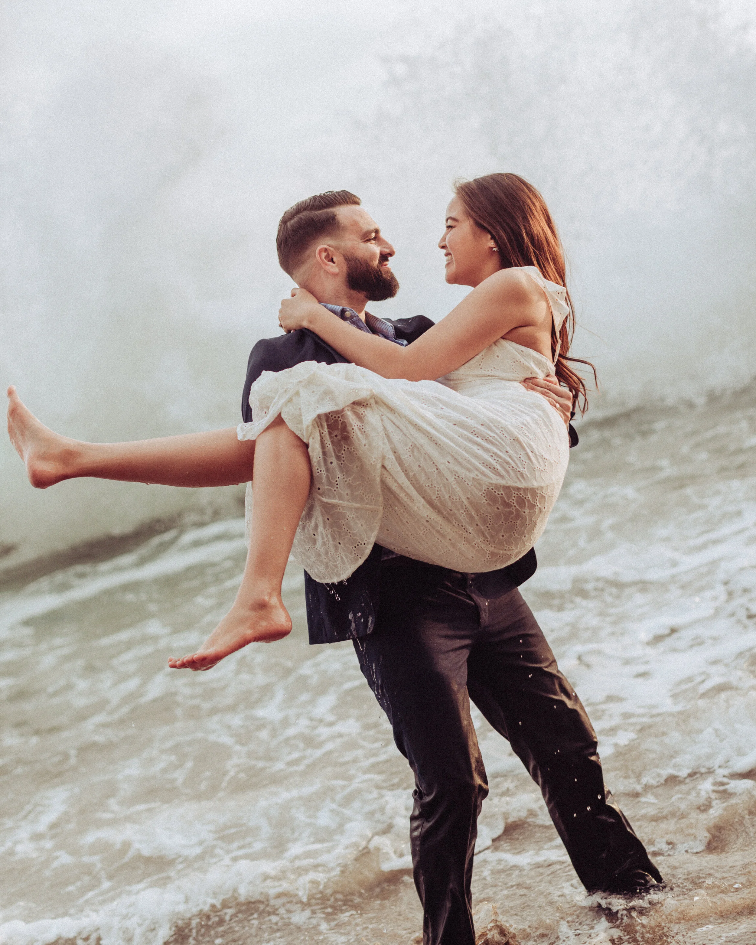 A happy couple at the beach. The man is holding the woman in his arms, they are looking at each other and smiling. The woman is wearing a light dress, and the man is wearing dark pants and a jacket. The background shows the ocean and cloudy sky.