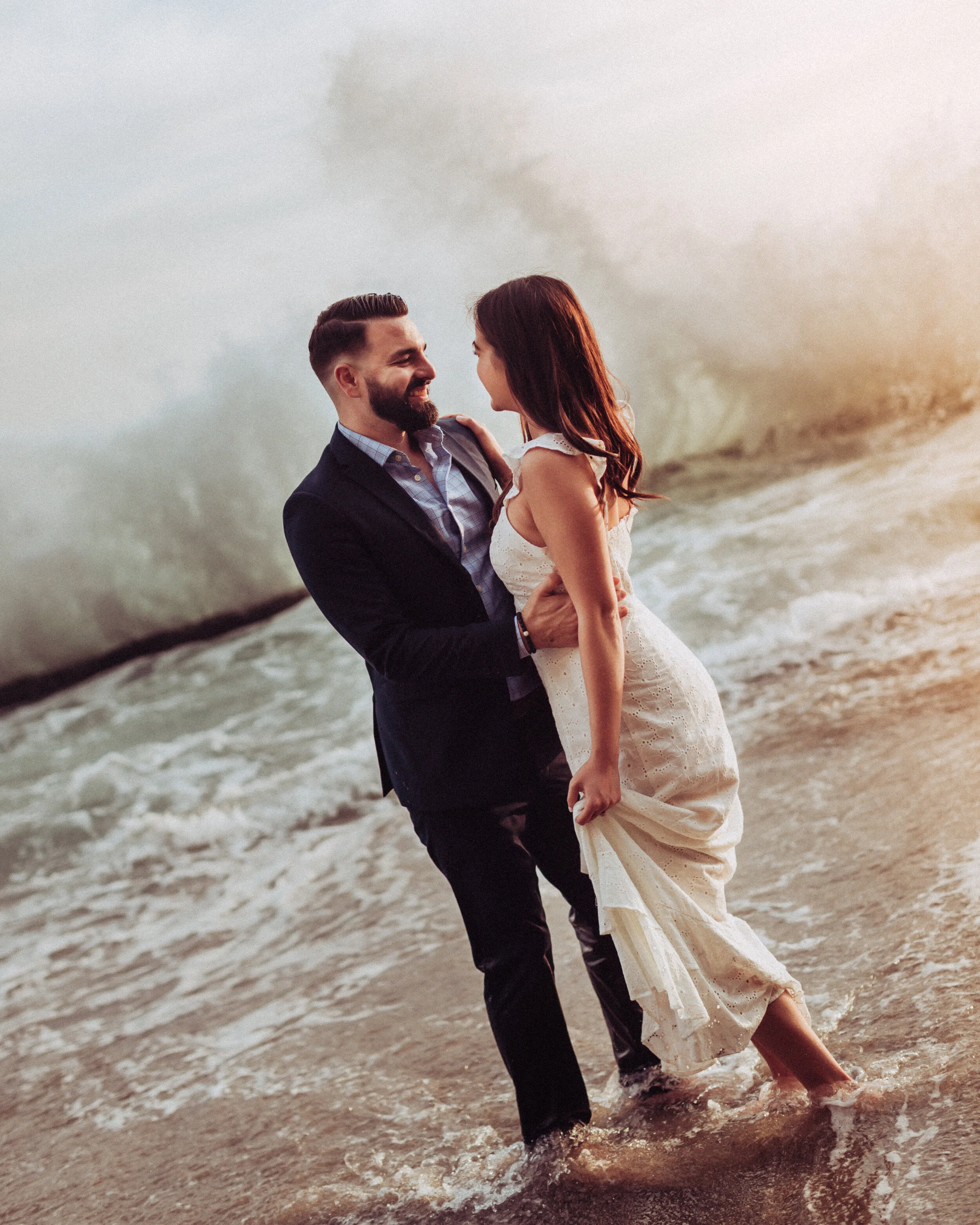 A man in a suit and a woman in a white dress are standing in the water on the beach, smiling and looking at each other, with waves crashing behind them at sunset.