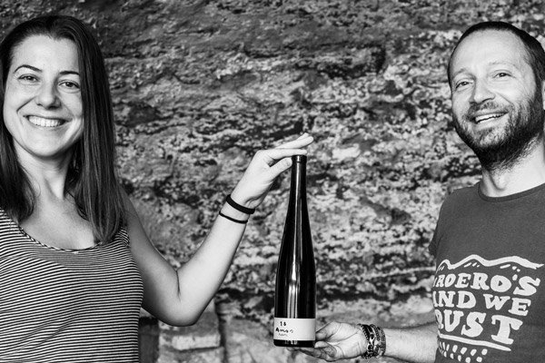 Two smiling people, a woman and a man, holding a bottle between them outdoors against a rocky background.