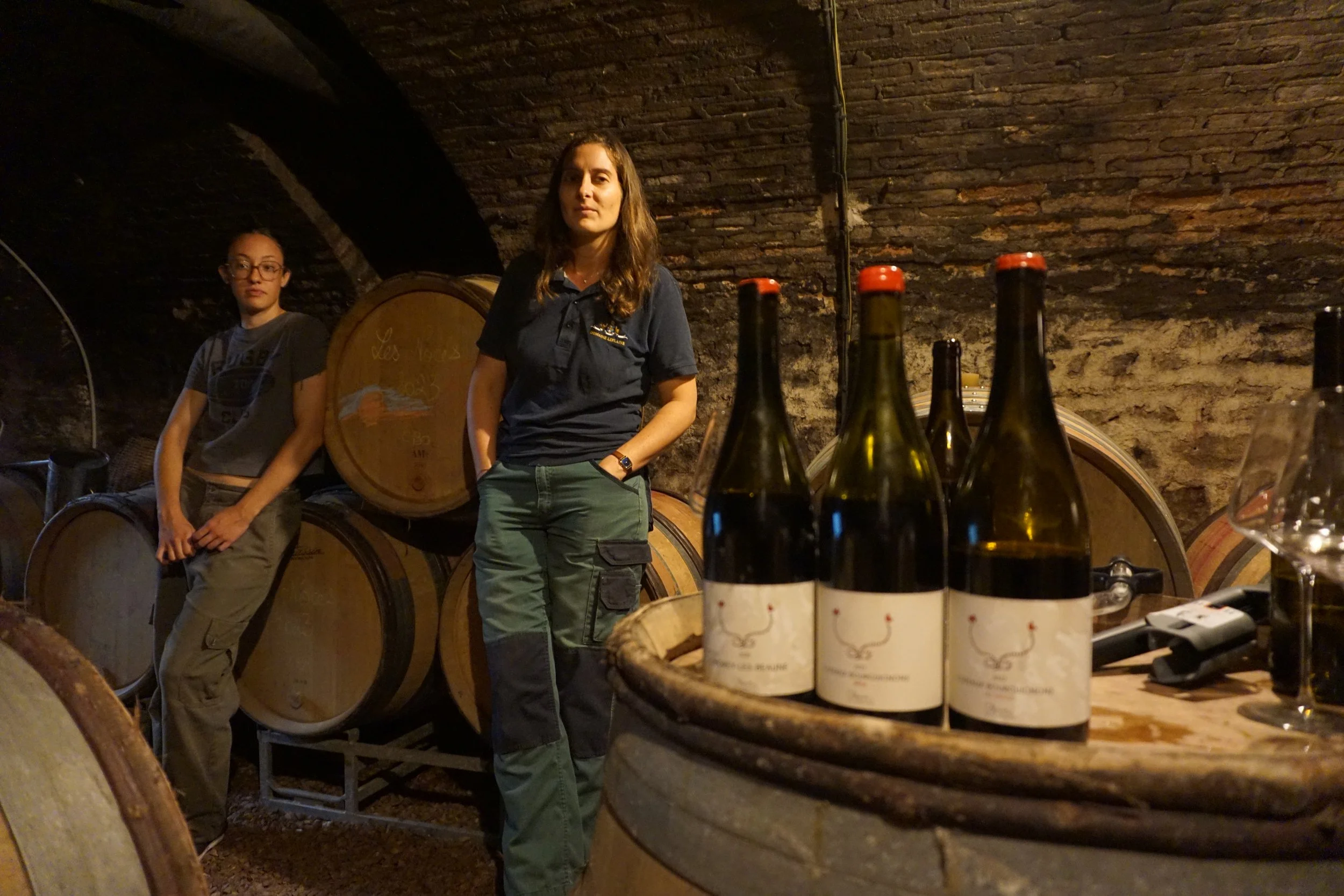 Two women standing in a wine cellar with wine barrels and bottles, one woman wearing a black polo shirt and green pants, the other in a gray T-shirt and beige pants.