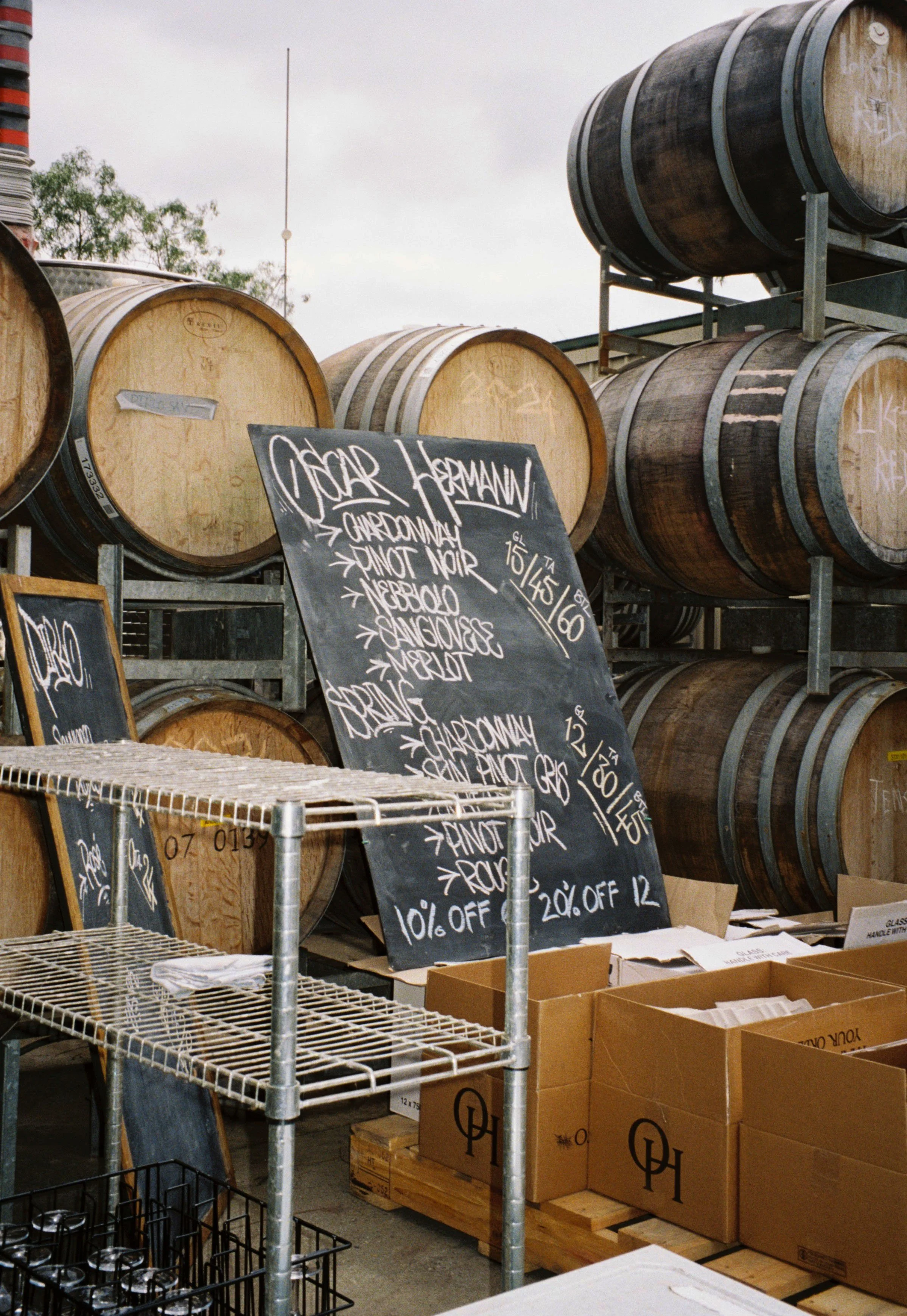 A wine storage area with wooden barrels stacked on racks, chalkboard menus displaying wine options, and cardboard boxes on pallets.