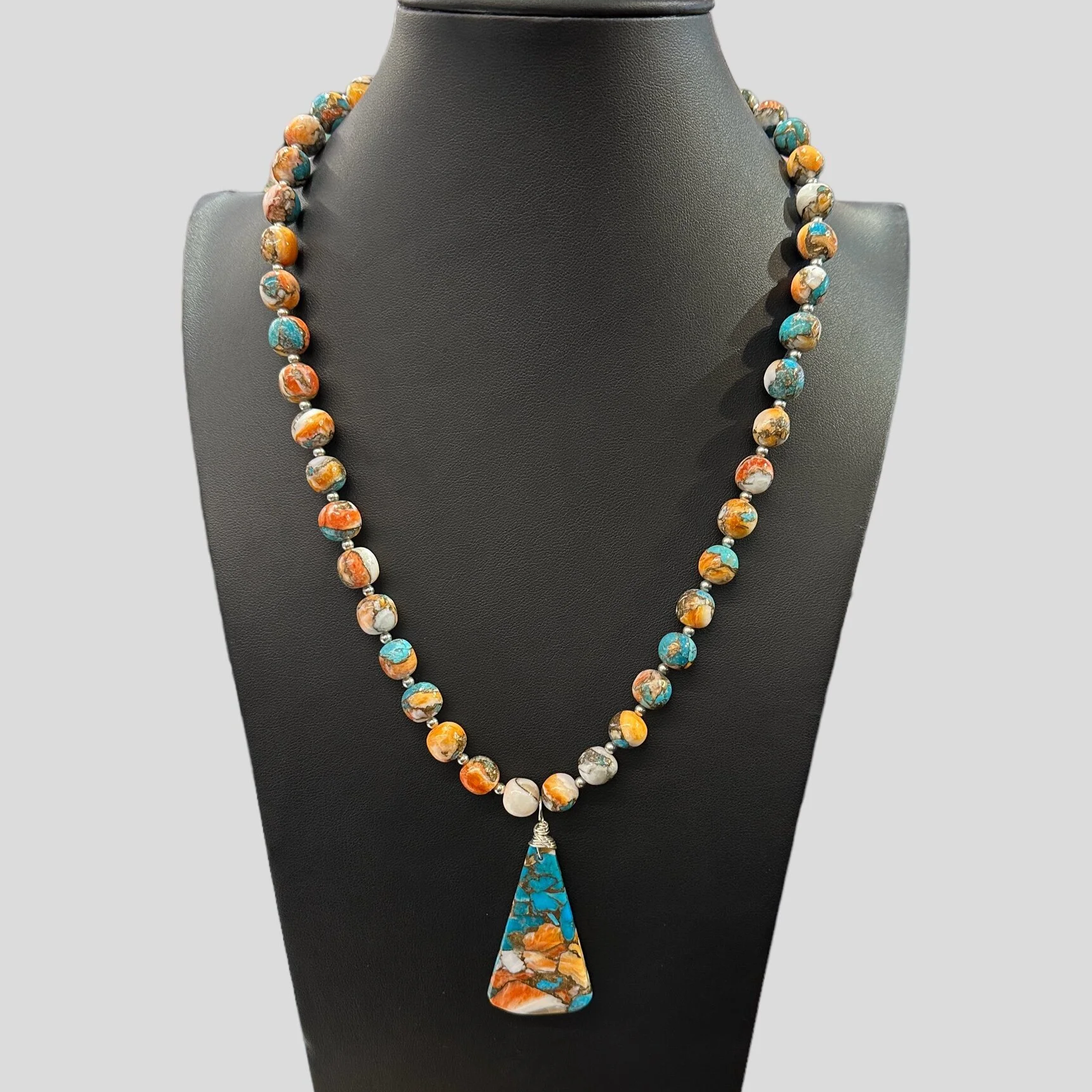 Beaded necklace with a large, colorful turquoise and orange stone pendant displayed on a black jewelry stand. The necklace features small round beads with multi-colored patterns.