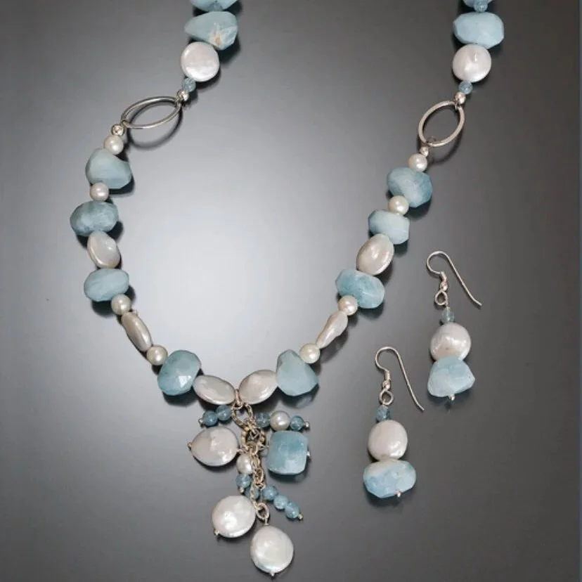 A necklace and matching earrings made of white pearls, light blue irregularly shaped beads, and silver accents on a dark surface.