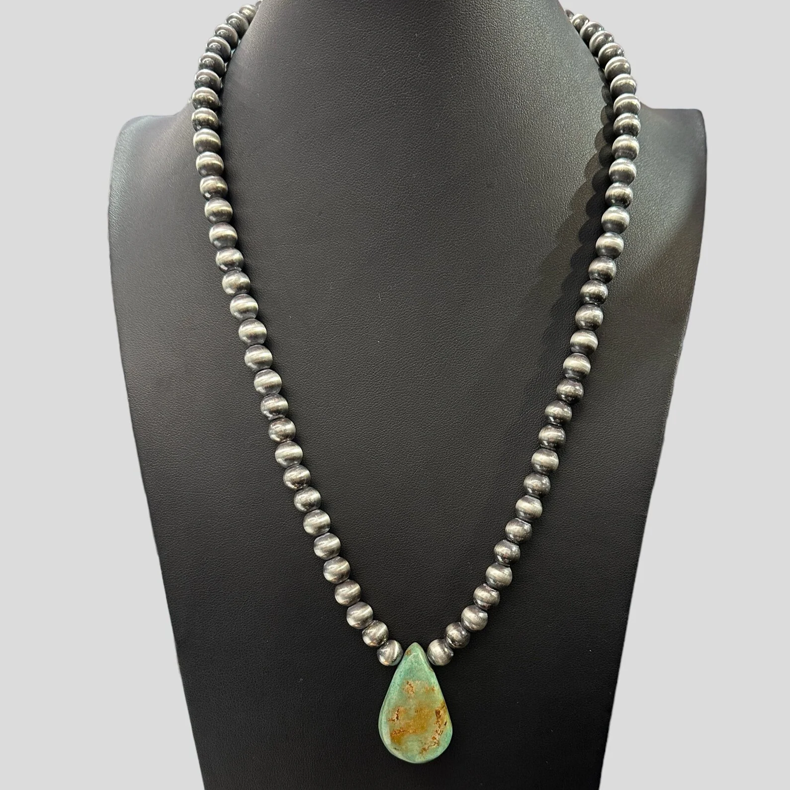 A necklace with silver beads and a teardrop-shaped green pendant with rust-colored spots on a black display bust. The text at the bottom reads, "Victoria's Southwest Designs."