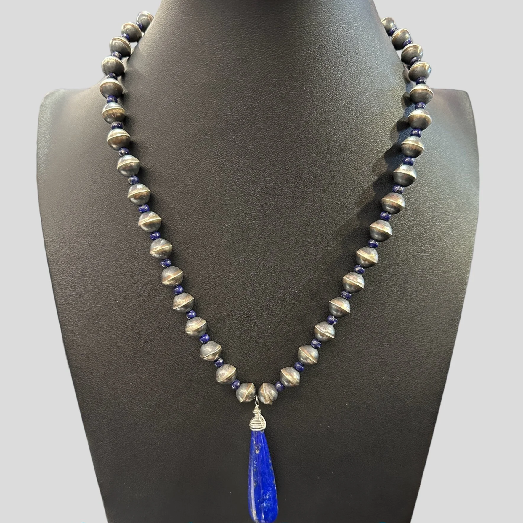 A necklace with round metallic beads and a blue teardrop pendant displayed on a black jewelry stand. Text overlay reads "Victoria's Southwest Designs."