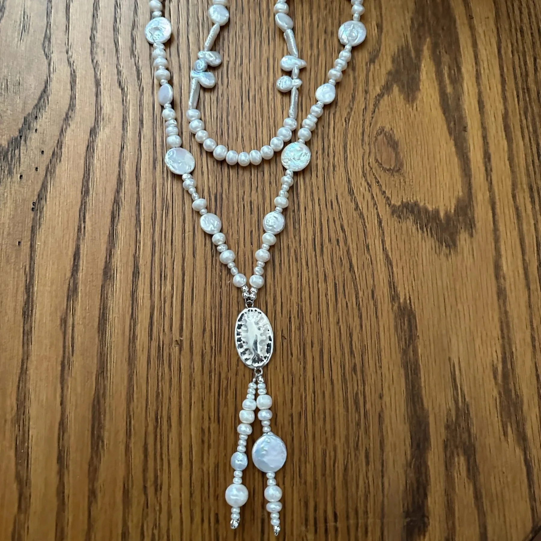 A white pearl necklace with a silver pendant and hanging pearl drops, laid out on a wooden surface. Text at the bottom reads "Victoria's Southwest Designs" in blue.