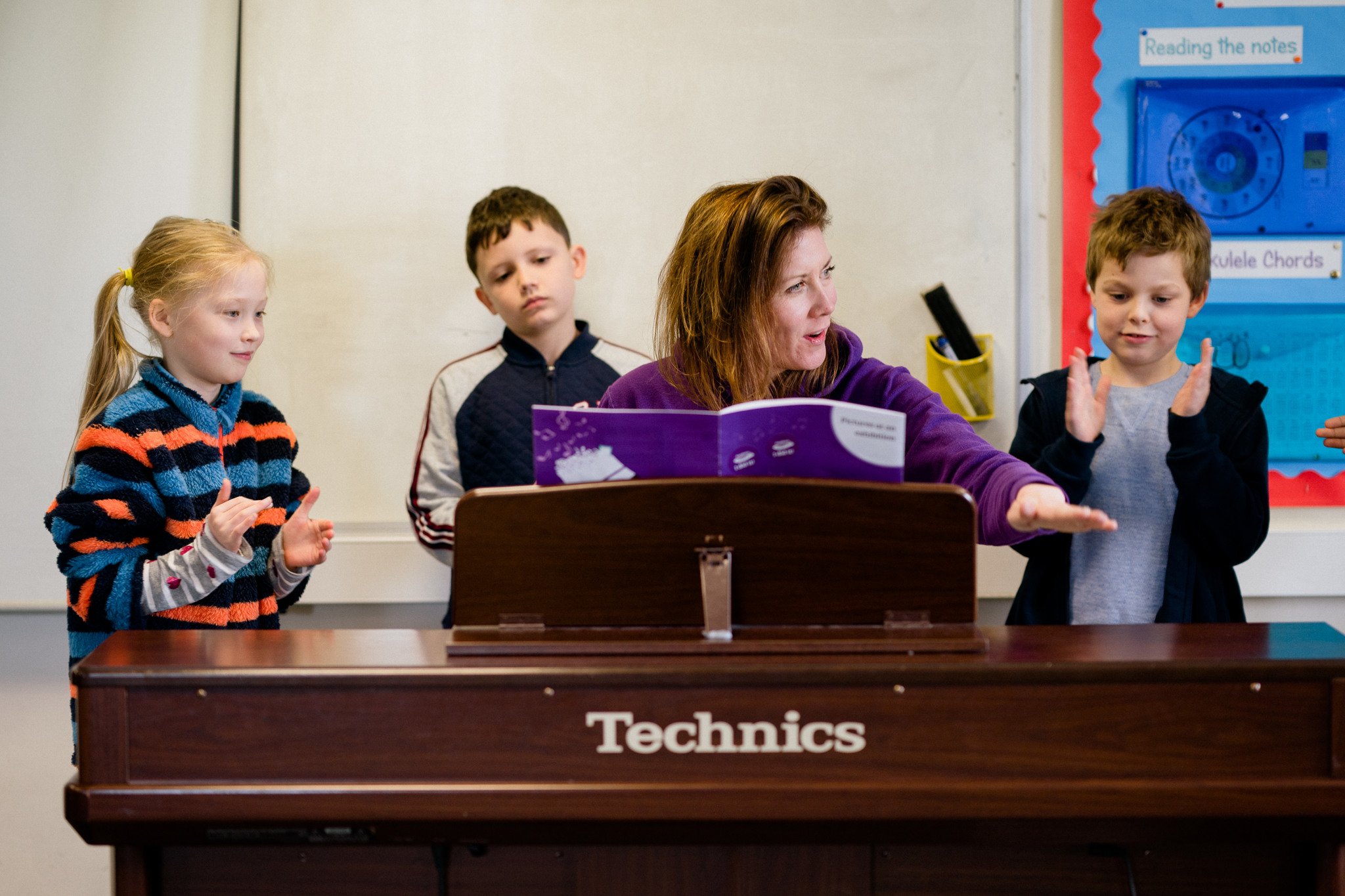Children gather around a piano teacher at her piano, clapping to the rhythm.