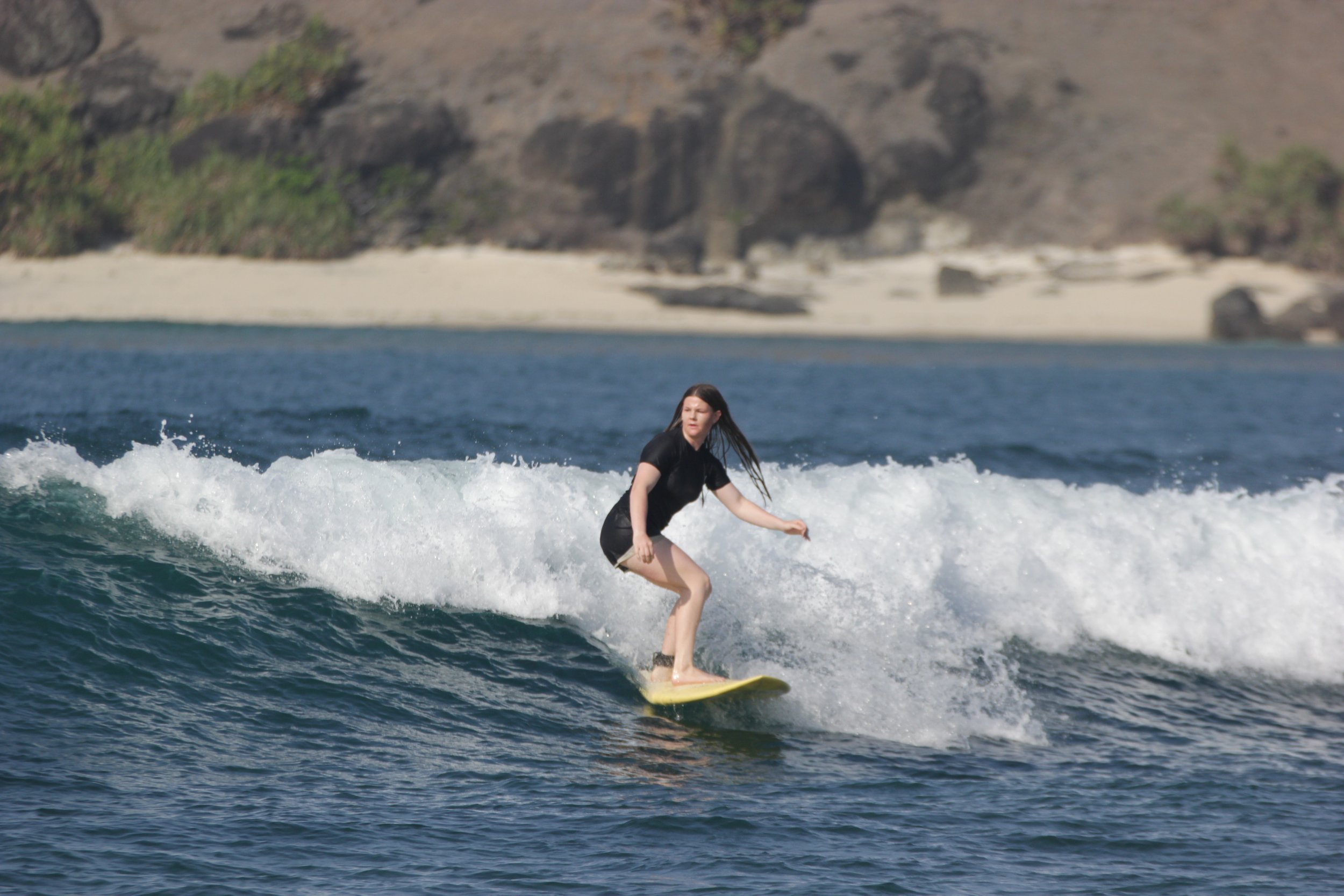 A woman surfing on a yellow surfboard on the ocean with cliffs in the background.