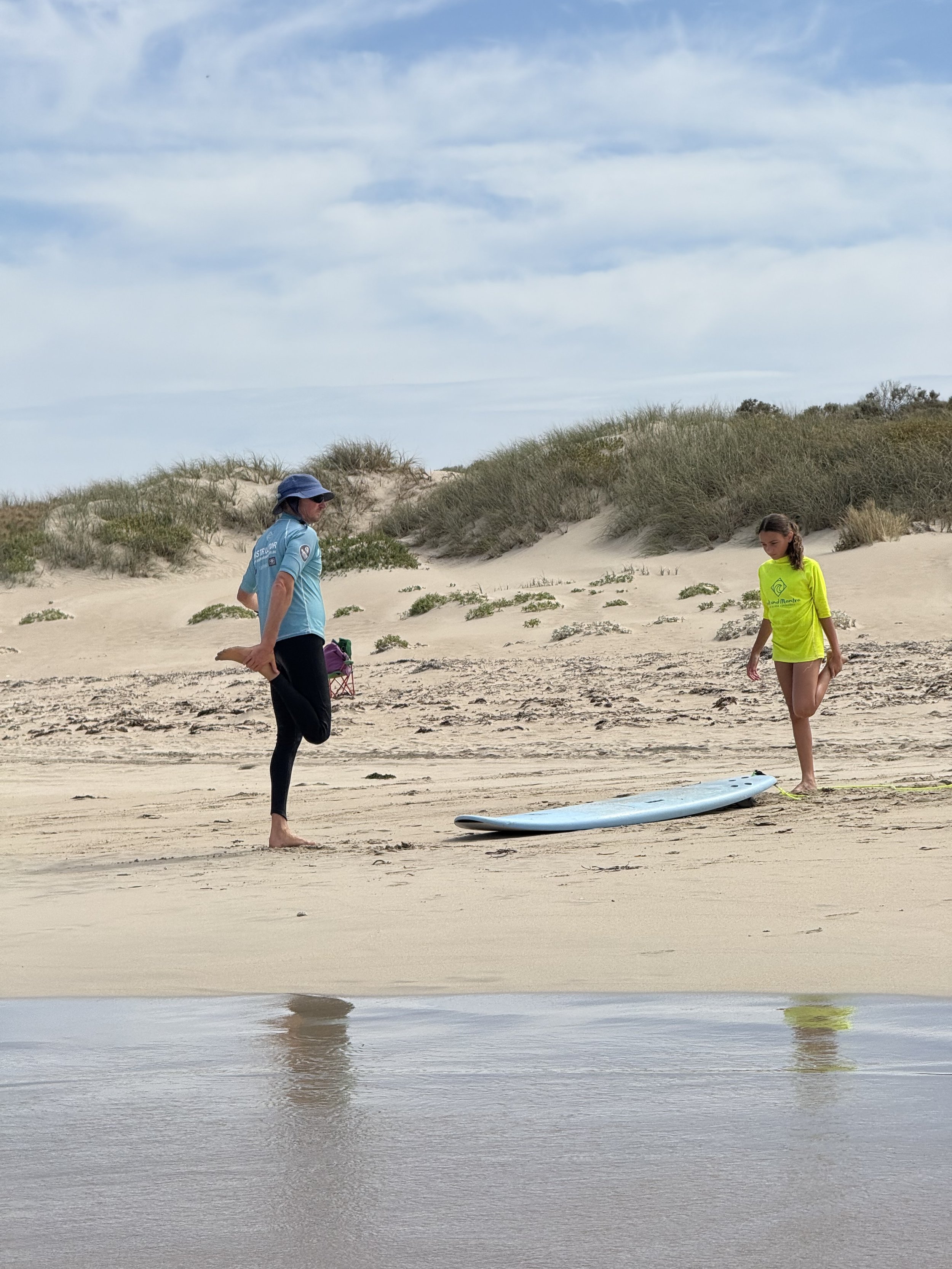 A man and a young girl on a sandy beach stretching before they go surfing. There are sand dunes and shrubs in the background under a partly cloudy sky.