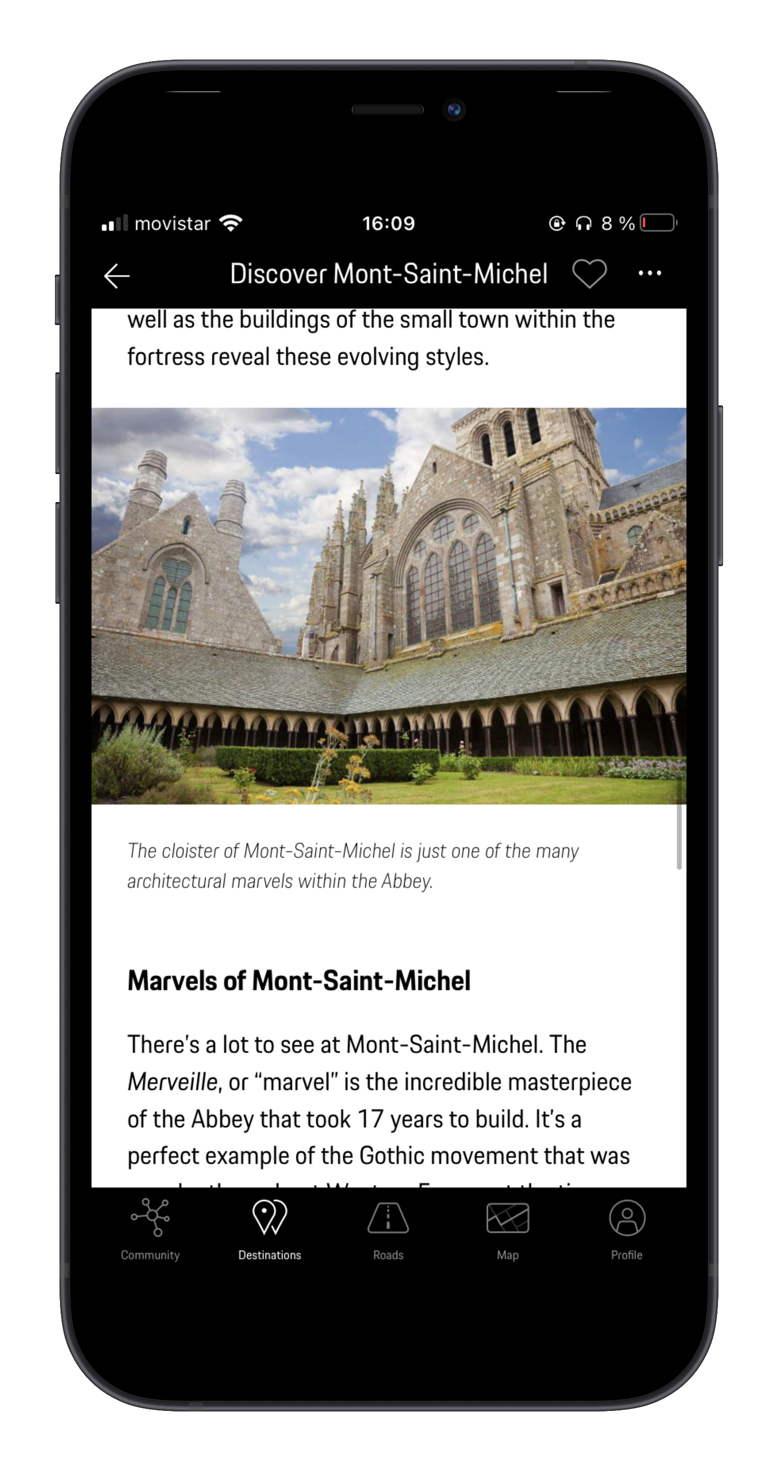 Mobile phone displaying an article about Mont-Saint-Michel, showing a photo of the abbey with Gothic architecture, green gardens, and a partly cloudy sky.
