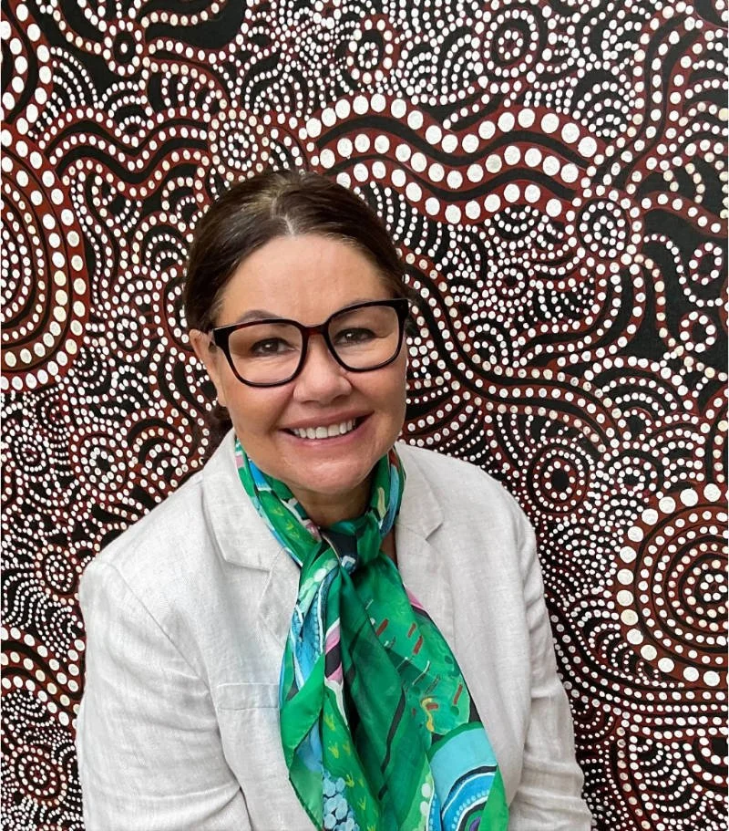 Maria Watson-Trudgett, wearing green scarf nad blazer, smiling in front of a detailed dotted pattern background - which is Maria's artwork.