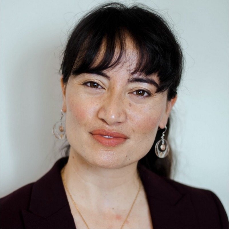 Close-up portrait of a woman with dark hair, wearing earrings and a dark blazer, smiling softly against a plain background.