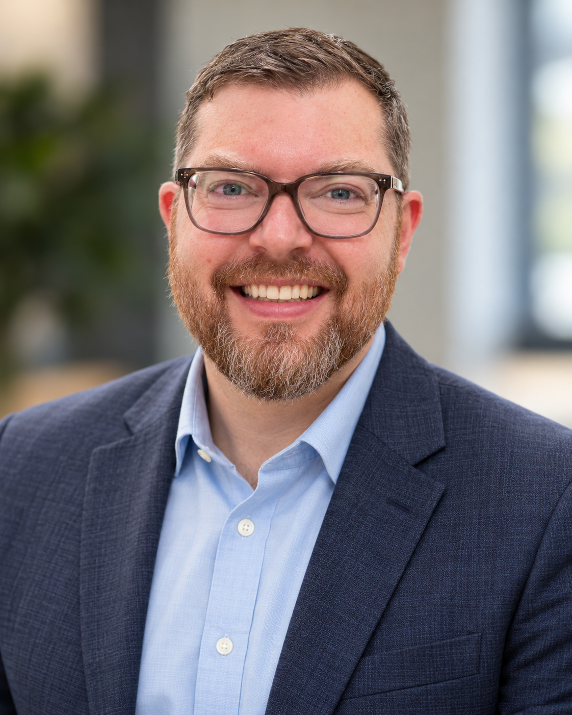 Close-up of a man's face with curly brown hair, glasses, and a beard, smiling while wearing a gray suit and blue striped tie.