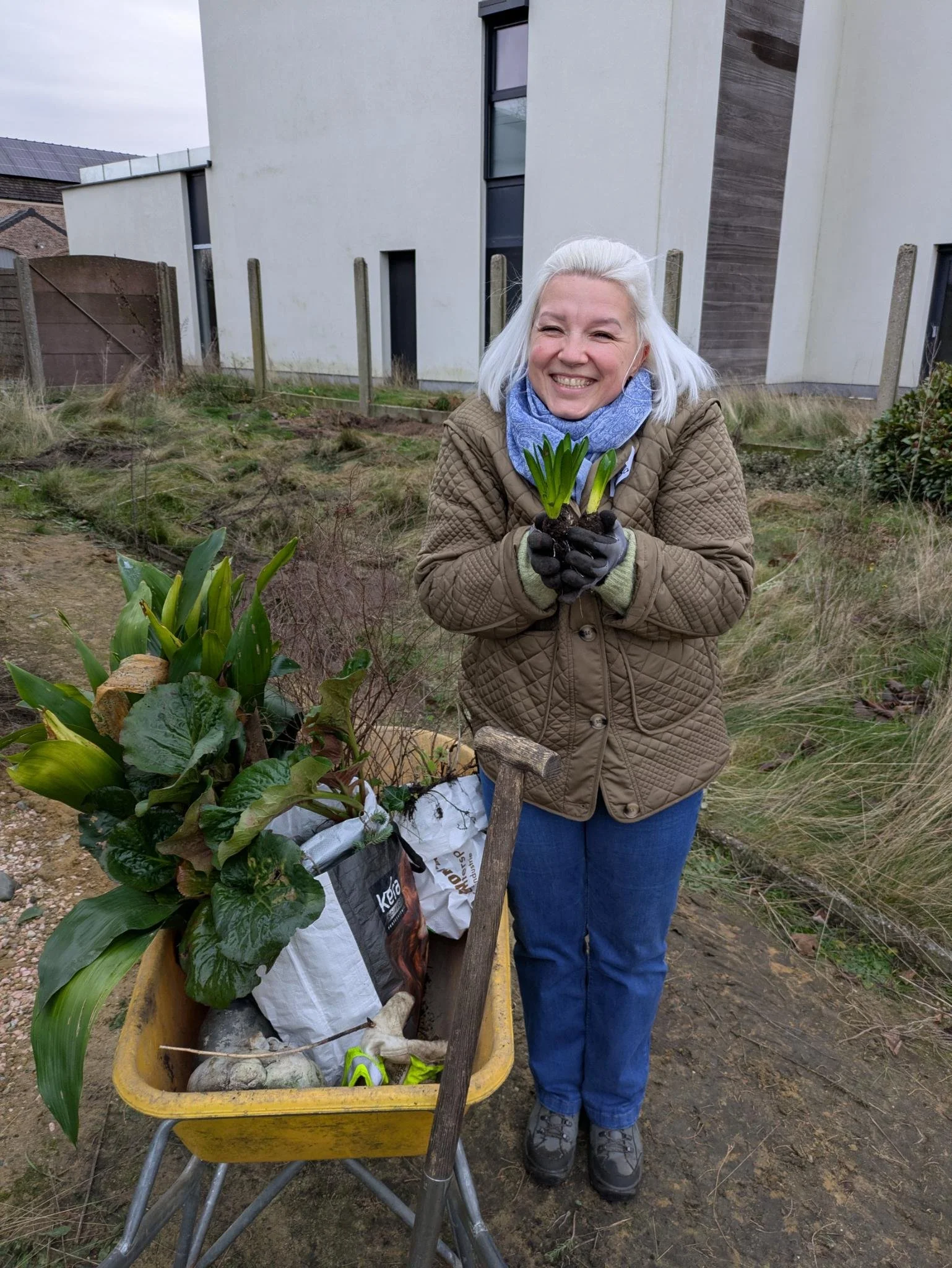 Vriendelijke vrouw met wit haar, gekleed in een beige jas en blauwe broek, glimlachend in een tuin, terwijl ze een tulp in haar handen vasthoudt.