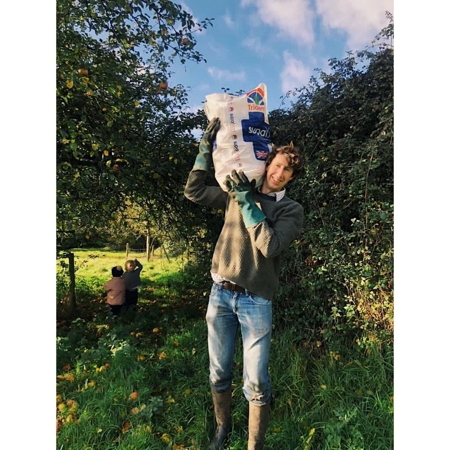 Young man in overalls and gloves lifting a large bag of apples in an orchard with two children in the background near trees and a blue sky.