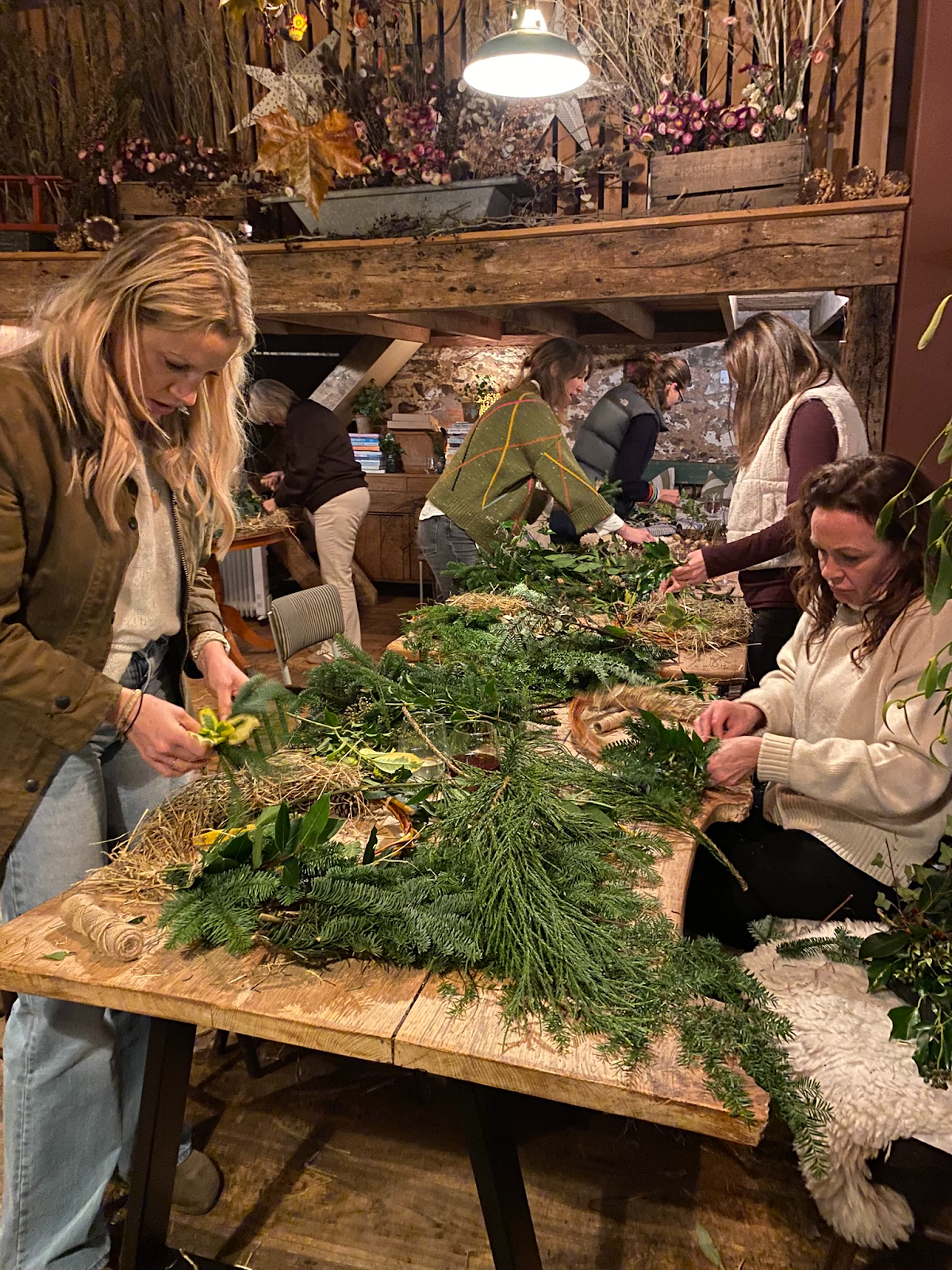 Person arranging a variety of green foliage and sprigs on a wooden table, likely creating a floral or plant arrangement.