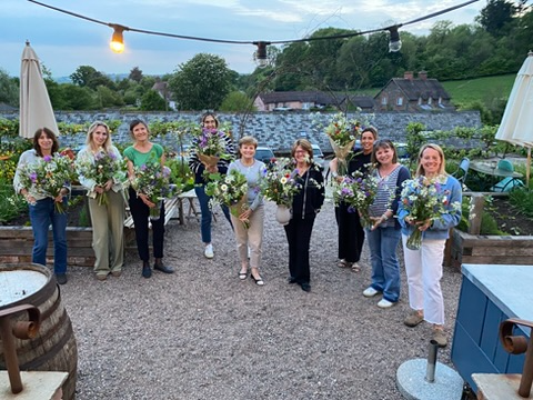 A large white vase with pink roses and various other colorful flowers, placed on a tall gray pedestal against a textured beige wall.
