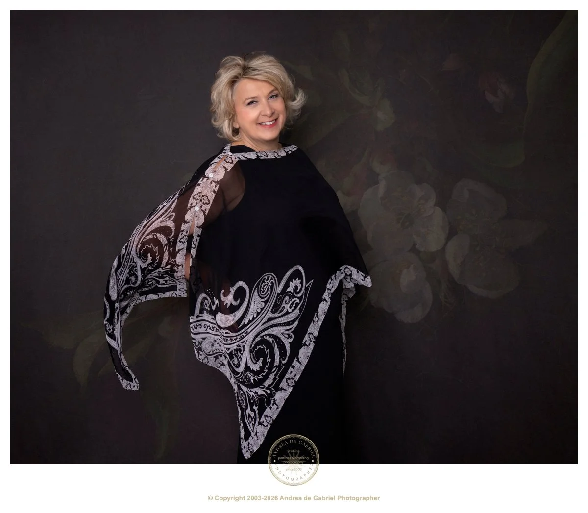 mature woman with blonde hair, wearing a flowing clack sheer cape, with white details on the edge, smiles at the camera