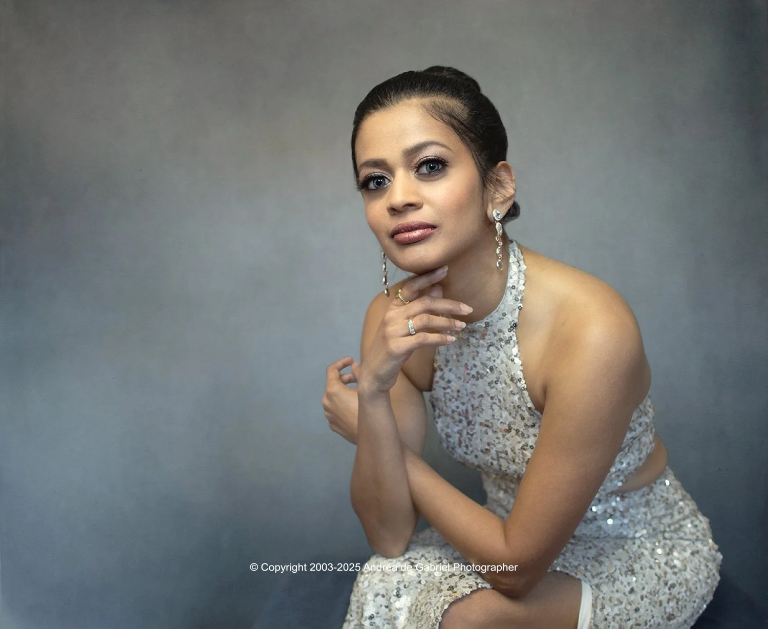 A woman in a sleeveless, sequined dress with earrings and rings, posed with her hand on her chin against a plain gray background.