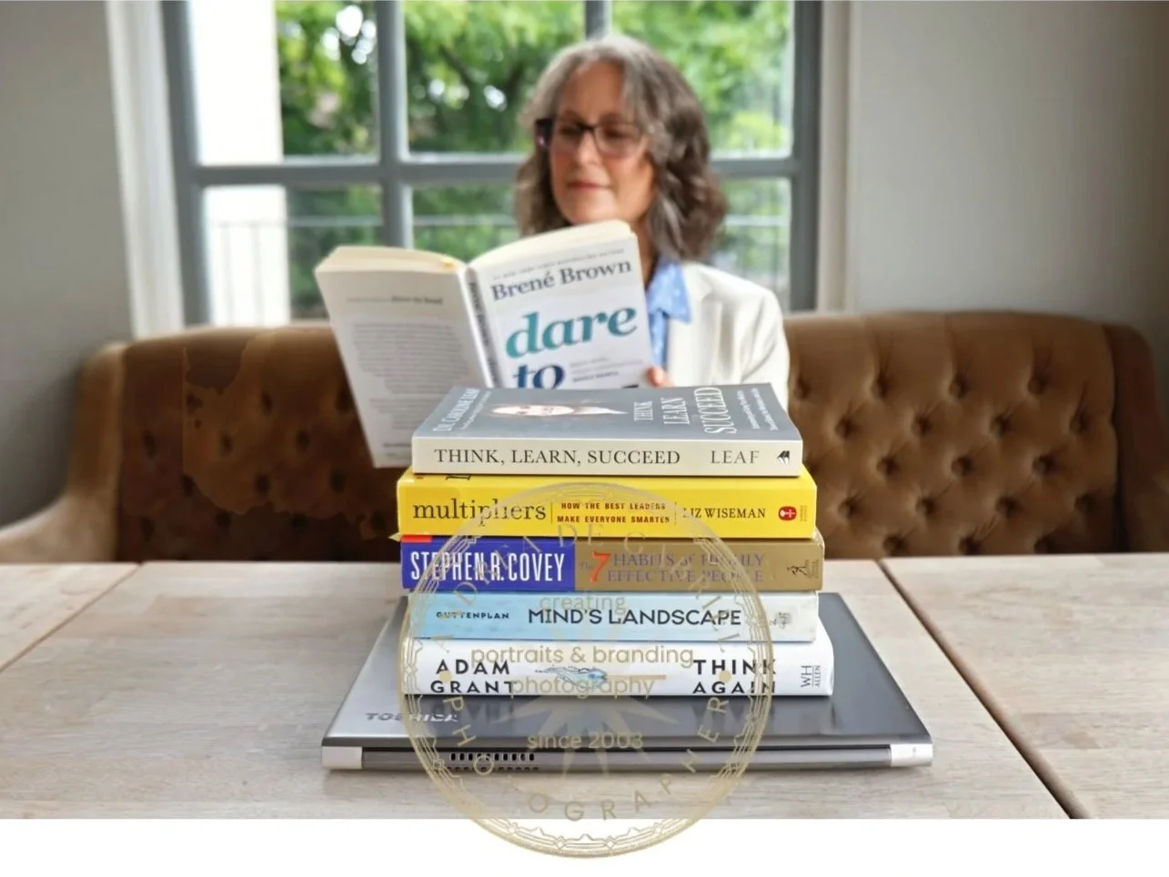 woman in white jacket reads a book behind a pile of other books