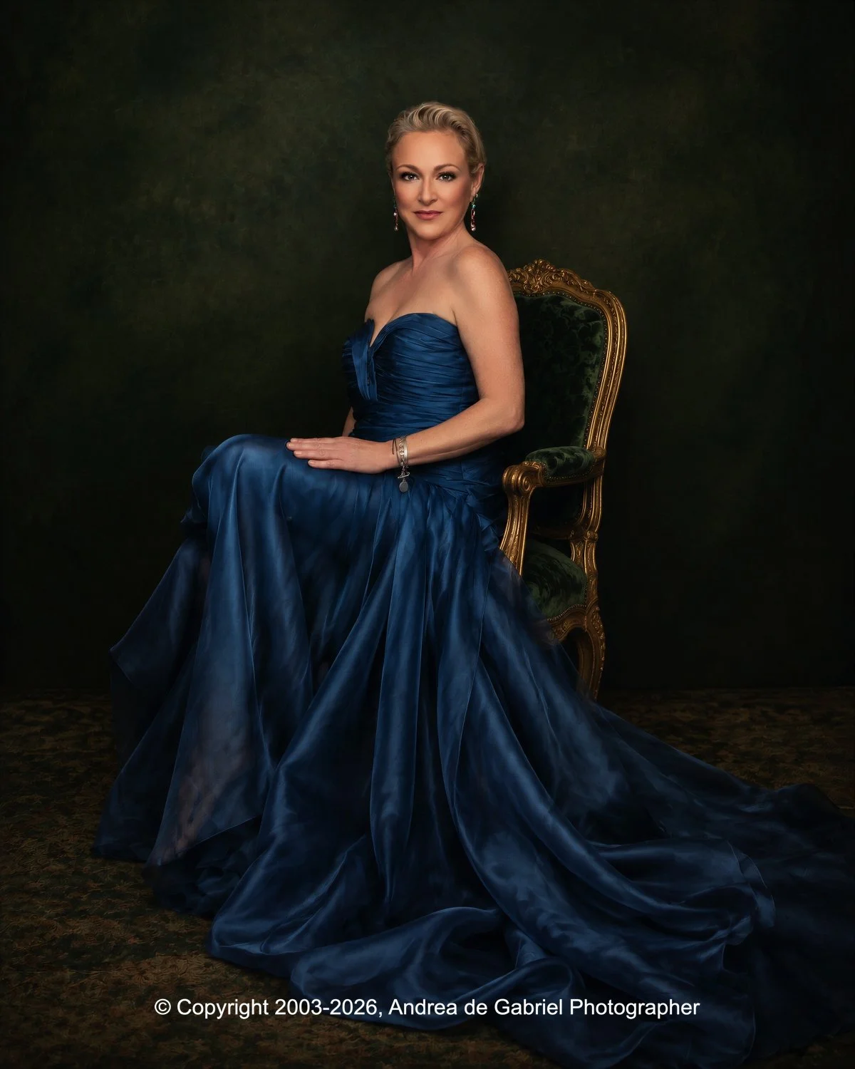 A woman in a strapless, royal blue gown sitting on an ornate, green velvet and gold chair against a dark background, looking at the camera.