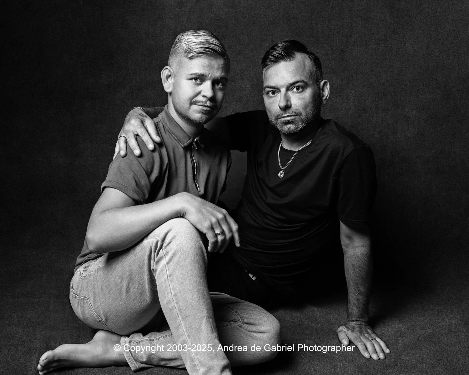 Cornwall Portraits same sex couple portraits. Black and white photo of two men sitting on the floor, one with his arm around the other's shoulder, both looking at the camera.
