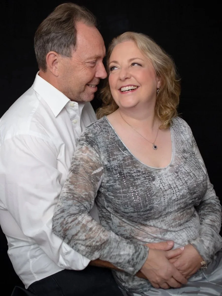 A middle-aged man and woman smiling and cuddling against a solid black background, with the man leaning close to the woman and the woman smiling joyfully.