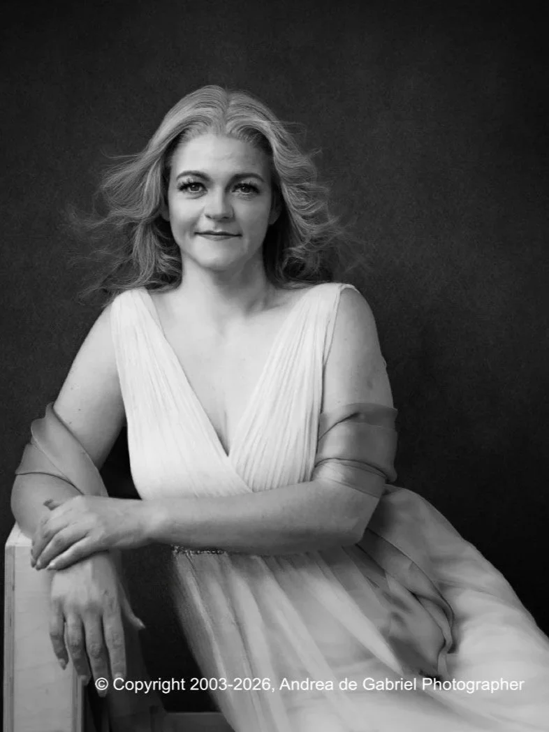 Black and white portrait of a woman with shoulder-length wavy hair, sitting and looking at the camera, wearing a sleeveless, pleated dress with a deep V-neck, and a sheer shawl or wrap on her arms.