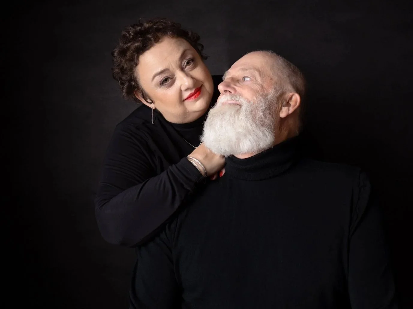 A woman with short curly hair and red lipstick holds an older man with a white beard wearing a black turtleneck, posing against a black background.