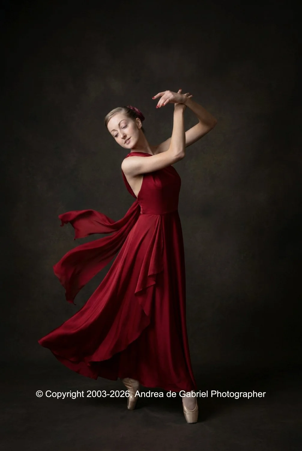A woman in a flowing red dress dancing gracefully on pointe with her arms raised, against a dark background.