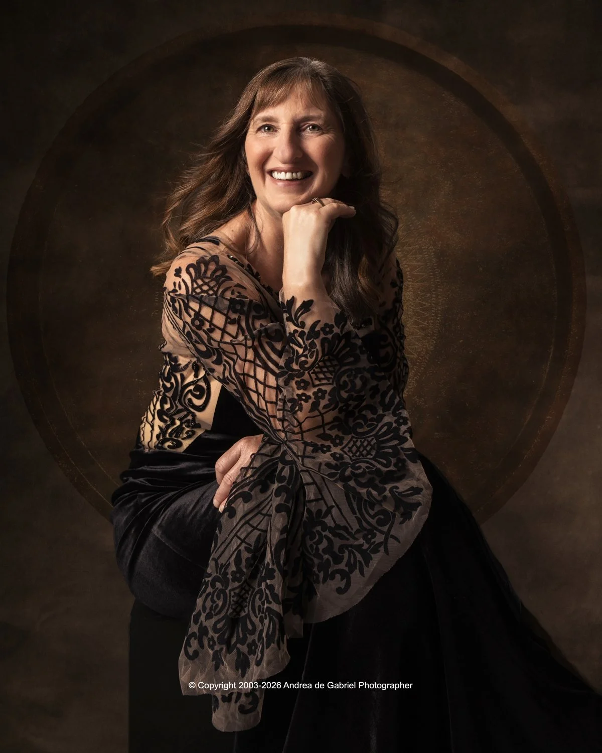A woman with long brown hair wears a black and beige patterned lace top and a black skirt, smiling and posing with her right hand under her chin against a dark, textured circular background.