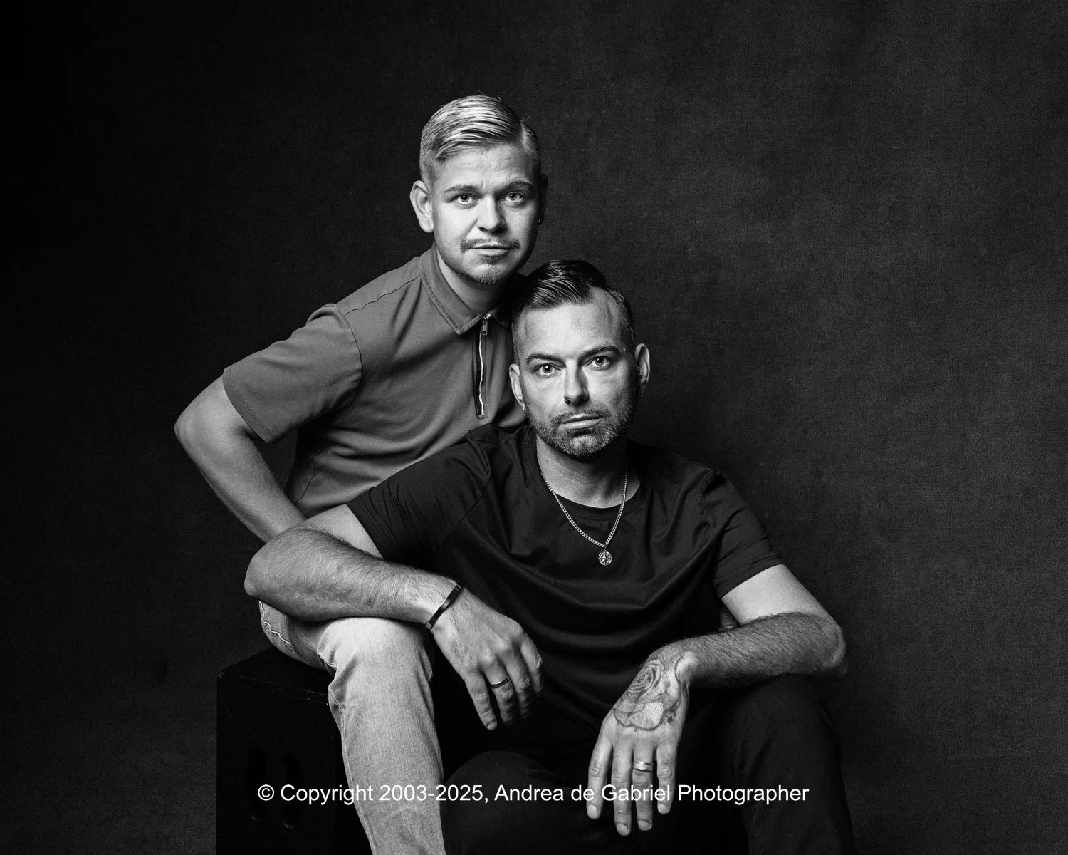 A same sex couple pose for a black-and-white portrait against a dark background, one sitting with arms crossed and the other standing behind him with a hand on his shoulder.