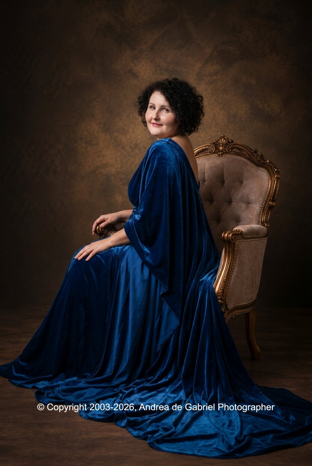 A woman with curly black hair sitting on a vintage upholstered chair, wearing a flowing blue velvet gown, against a dark, textured background.