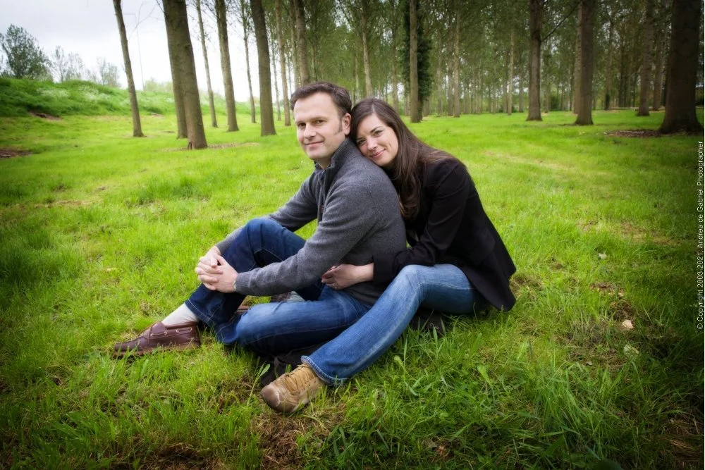 A couple sitting on grass in a wooded park, with the woman resting her head on the man's shoulder and both smiling.