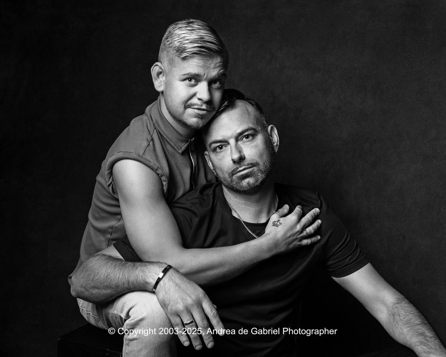 Same sex couple sitting together, one resting his arm on the other's shoulder, against a dark background. Black and white photo.