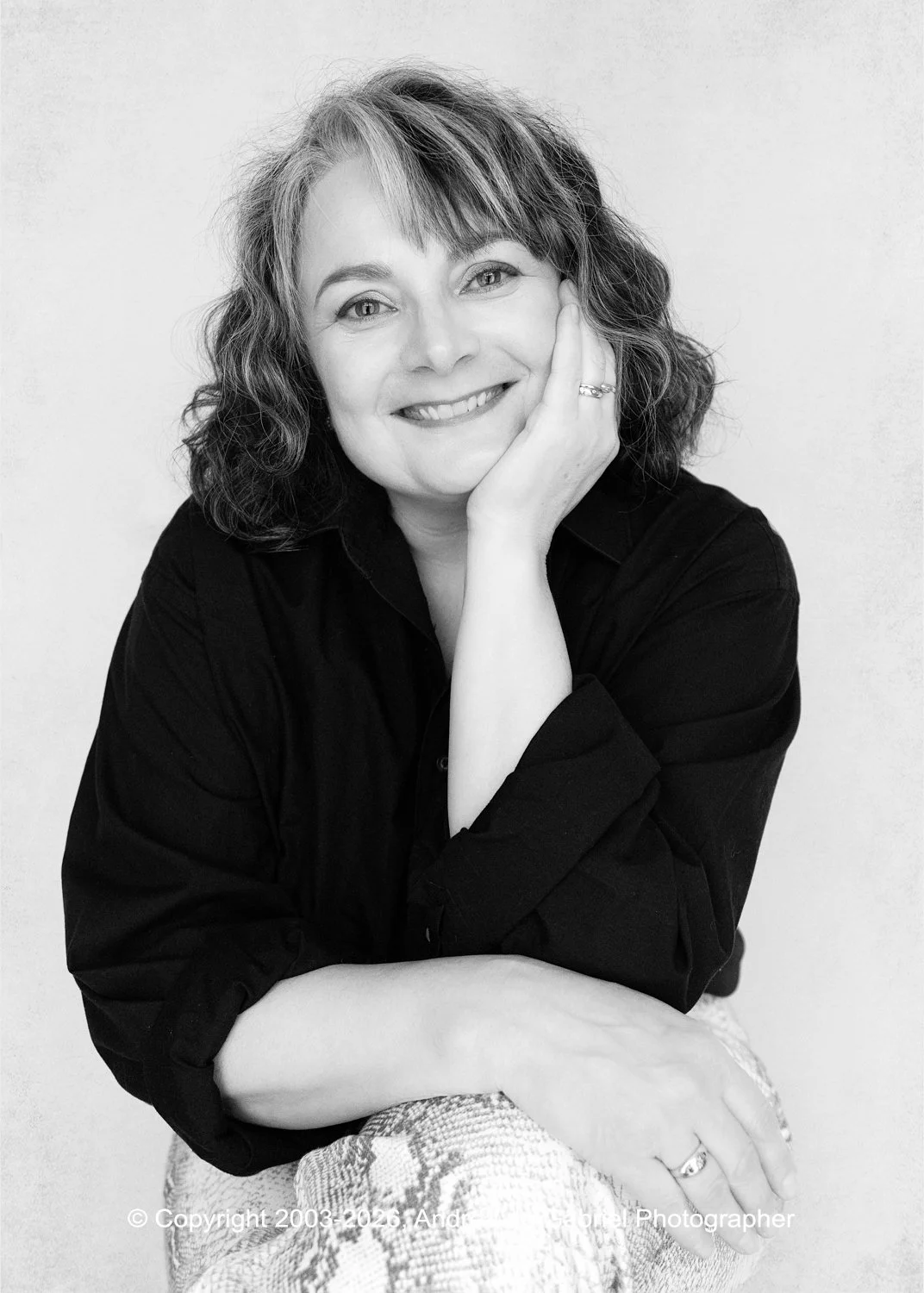 Black and white portrait of a woman with shoulder-length wavy hair, smiling, wearing a black shirt, resting her face on her hand.