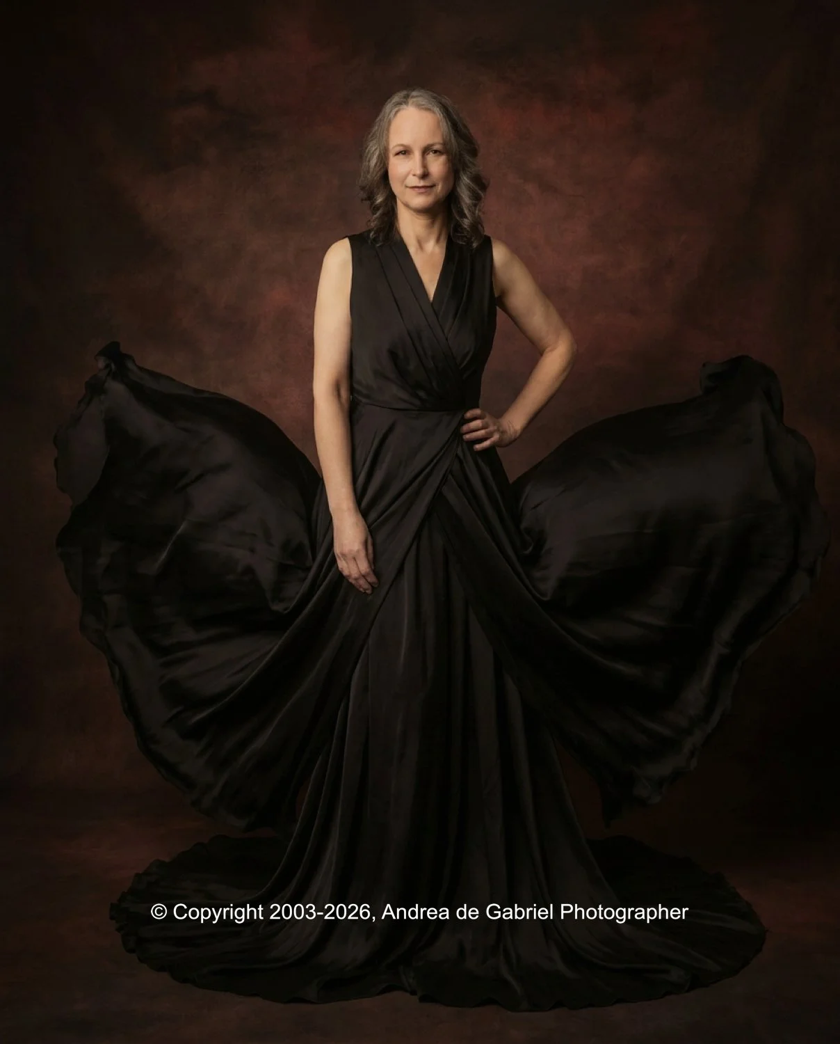 A woman in a flowing black gown standing with hand on hip against a dark, textured background.