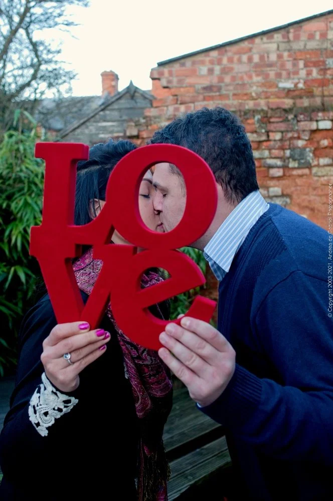 A couple is kissing behind a red cutout of the word "LOVE," which they are holding in front of their faces, outdoors with a brick wall and trees in the background.