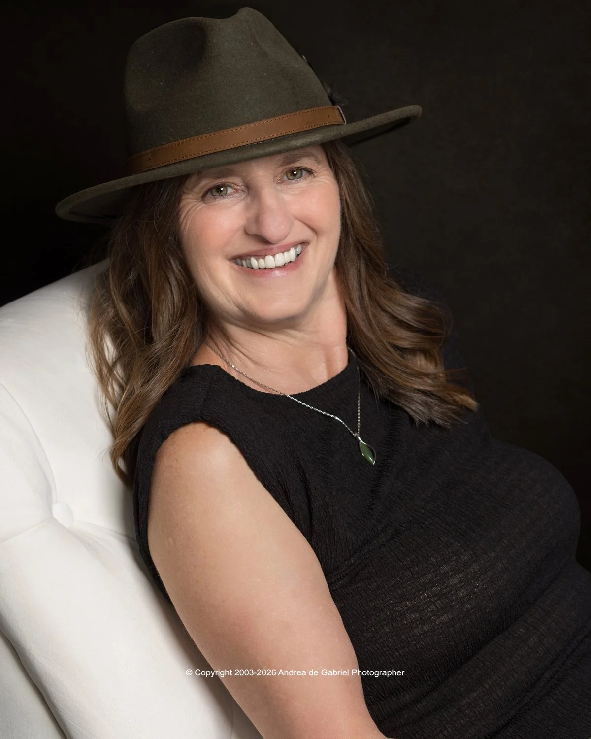 A woman wearing a large hat with a brown band, smiling while sitting on a white chair, dressed in a black top with a silver necklace featuring a green pendant, against a dark background.