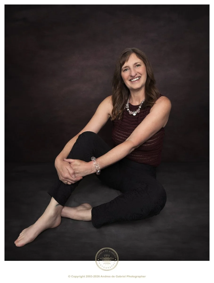 mature woman sits on the ground, holding her knee. she has mid length brown hair, and is smiling for the camera. she is wearing black trousers and a maroon top.