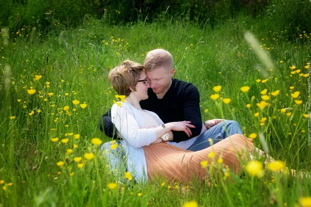A couple sitting together on a grassy field filled with yellow flowers, sharing an intimate moment with their foreheads touching and eyes closed.