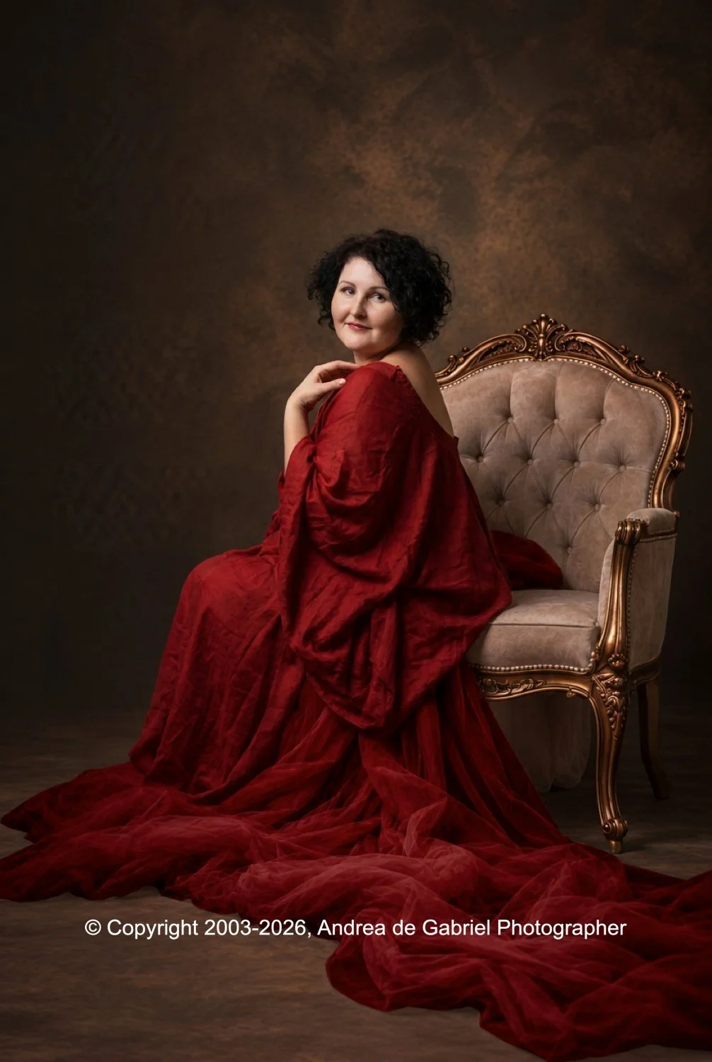 A woman with dark curly hair wearing a long red dress seated on an ornate vintage chair in a studio setting with a dark textured background.